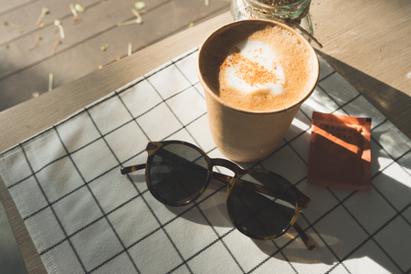 cappuccino in take away coffee cup on table cloth with dry flower,sunglasses on wood table with sunlight hard shadow at window in evening.food and drink lifestyle leisure conceptの写真素材