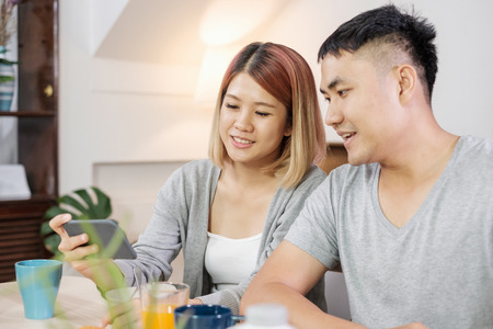 happy asian couple in pajamas sitting at table in kitchen and watching video on mobile togerther at home in morning and having cereal breakfastの写真素材