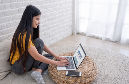 Asian female sit with knees up on floor using laptop see social media website on wicker stand near window in living room at home.Work at home concept.work from home.relax lifestyleの写真素材