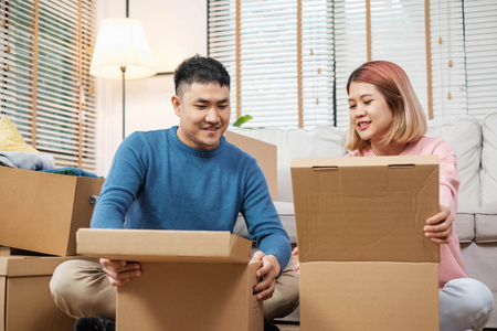 Happy asian couple open cardboard boxes  while moving to new home at sofa in living room.unpacking for new houseの写真素材