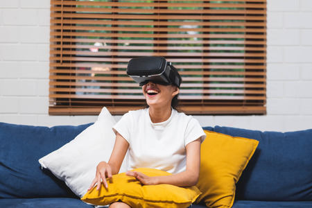 Asian woman wear vr headset glasses and play virtural reality  sitting on sofa at home.digital tehcnology lifestyle.のイラスト素材