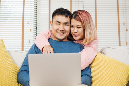 Asian couple watching video live on laptp on sofa in living room at home.lover spending time together.wife embrace husband from backの写真素材