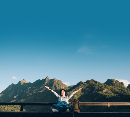 Asian woman traveler  sitting and arms up in the air at view point terrace at landscape view of mountain with cloud and blue sky in sunny day at forest.backpacker explore nature lifeの写真素材