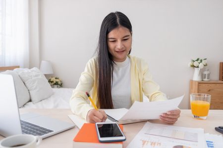 Asian female freelancer reading report on laptop and jot down note on table in bedroom at home.Work at home concept.work from home.relax lifestyleの写真素材