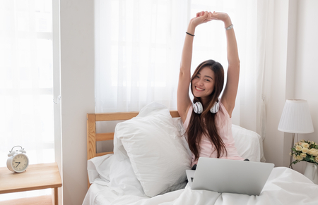Asian woman wear top tank raise arms up while using laptop on bed in bedroom in morning,relax lifestyle concept.looking at cameraの写真素材