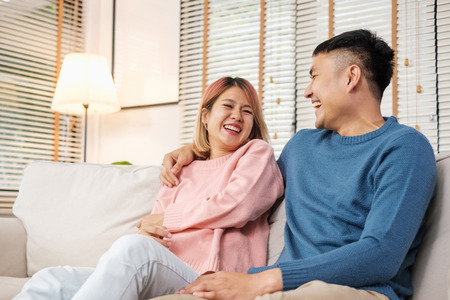 Asian couple embracing and laughing while sitting at sofa in house.lover couple in home conceptの写真素材