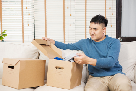 asian man open cardboard boxes  while moving to new home at sofa in living room.unpacking for new houseの写真素材