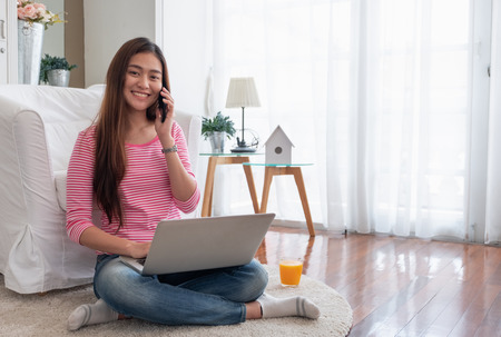 Happy asian woman talk on mobile and  typing laptop computer at home.female freelancer sitting on carpet on floor work at home in living room.の写真素材