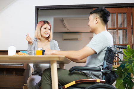 Asian couple breakfast at home.wife holding hand and encourage disabled husband sitting in wheelchair at kitchen table.couple in home conceptの写真素材