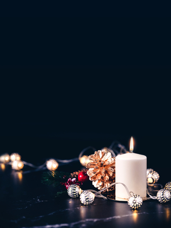 Christmas white candle with golden pine cone,mistletoe and light string glowing on black marble table with dark background.vertical banner holiday greeting card.の写真素材