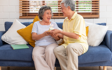 asian senior man surprise senior woman with birthday cake in living room at home.aging at home concept.の写真素材