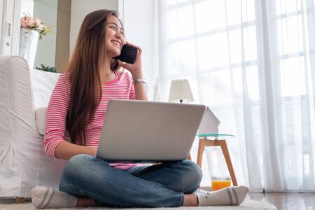 Happy asian woman talk on mobile and  typing laptop computer at home.female freelancer sitting on carpet on floor work at home in living room.の写真素材