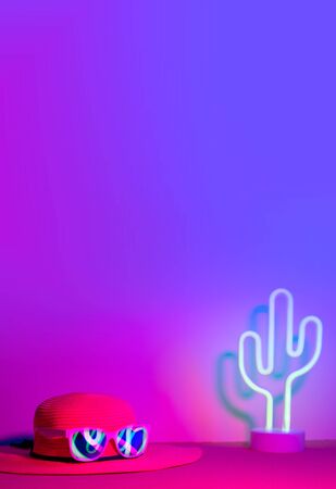 Summer with hat and sunglasses refection neon light with cactus on pink and blue light on table with copy space. vertical Trendy vacation holiday backgroundの写真素材