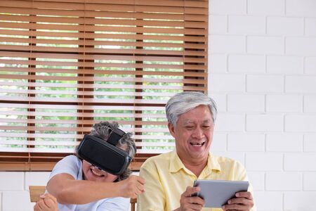 Asian senior couple play virtual realtiy glasses headset and tablet watching vr video and  have fun together in living room at home.senior with technology.aging at homeの写真素材