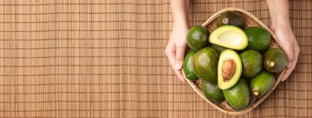 Hand holding organic avocados half cut with seed and whole fruit in basket on bamboo mat on table...