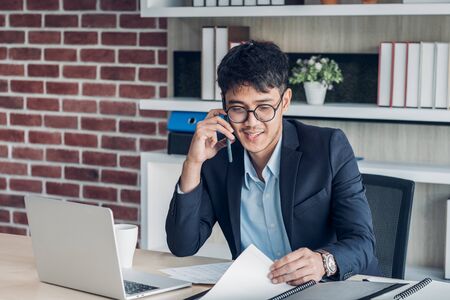 Young Asian businessman wear suit talking on mobile and see paperwork while working with laptop on desk table in modern office.connecting network online conceptの写真素材