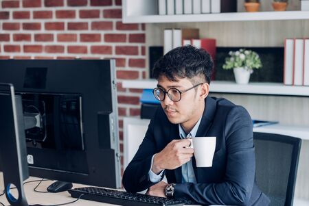 asian businessman wear suit working and drkink coffee when looking at desktop computer screen for online trade at table in modern office.の写真素材