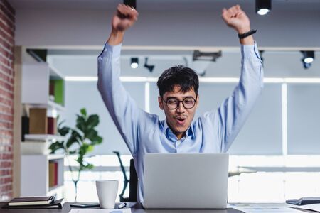 asian businessman arms up to celebrate success when job is done infornt of laptop on desk in modern office.の写真素材