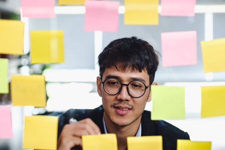 asian businessman looking and writing on sticky note for brainstorming ideas on clear whtieboard at modern office. analyze and synthesisの写真素材