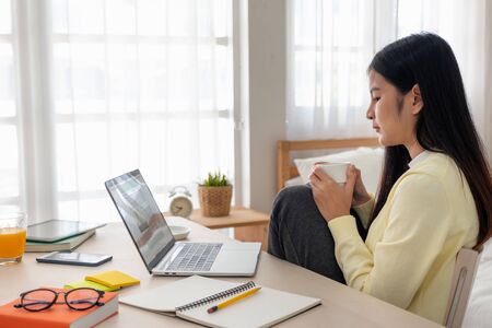 Asian female freelancer reading report on laptop and jot down note on table in bedroom at home.Work at home concept.work from home.relax lifestyleの写真素材