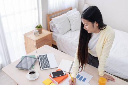 Asian female freelancer reading report on laptop on table in bedroom at home.Work at home concept.work from home.relax lifestyleの写真素材