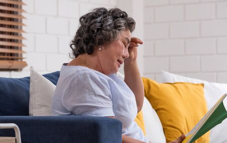 Asian senior woman sitting and reading book at sofa at home and  feeling headache ,high blood pressureの写真素材