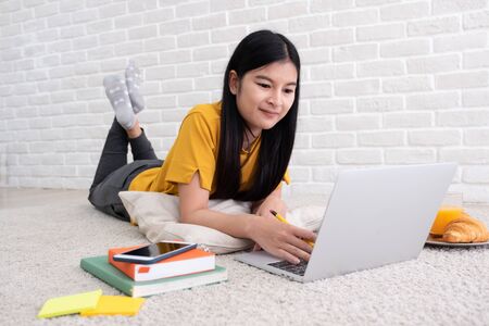 Asian female freelancer working on laptop computer at home.woman lying down on carpet at brick wall.working online lifestyleの写真素材