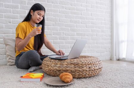 Asian female sit on floor using laptop on wicker stand and drink orange juice in living room at home with relax emotion.Work at home concept.work from home.relax lifestyleの写真素材
