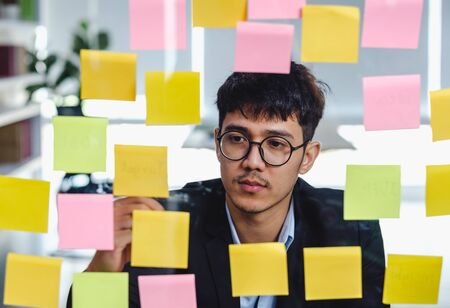 asian businessman looking and writing on sticky note for brainstorming ideas on clear whiteboard at modern office. analyze and synthesisの写真素材
