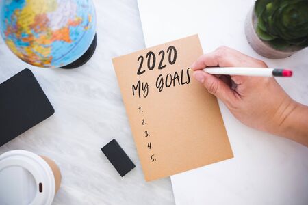 Hand holding pen writing 2020 my Goals on brown paper with blue globe,blackboard,coffee cup on...