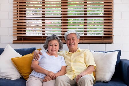Asian couple senior embracing and sitting comfortably on sofa and  watching tv in living room at home.の写真素材