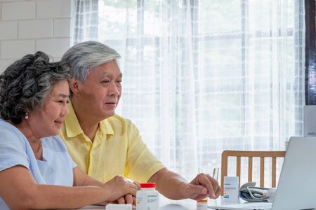 Asian couple seniors use laptop video conference with doctor about illness and prescription pill in living room at home.Healthy lifestyle.aging at home conceptの写真素材