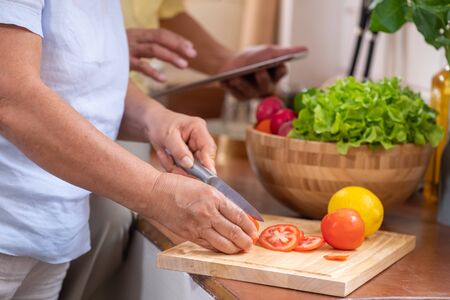 Asian senior wife slicing tomatoes on wooden cutting  board and using tablet computer to searching menu recipe.senior with technology lfiestyle.aging at homeの写真素材