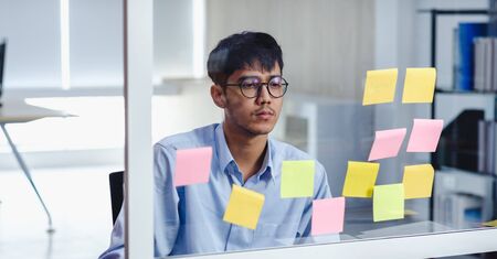 asian businessman looking and writing on sticky note for brainstorming ideas on clear whtieboard at...