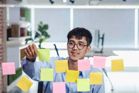 asian businessman looking and writing on sticky note for brainstorming ideas on clear whtieboard at modern office. analyze and synthesisの写真素材
