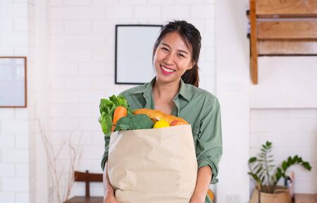 Happy asian woman  holding vegetable bag walking into kitchen after shopping at grocery store at home.vegan lifestyle conceptの写真素材