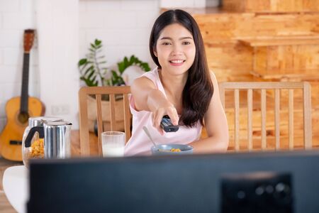 asian woman wathcing tv on demand and use remote control change television channel at breakfast in morningの写真素材