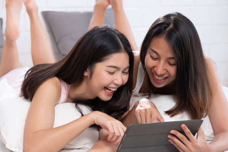 Asian lesbian couple lying down on bed using tablet browsing internet in morning at cozy homeの写真素材