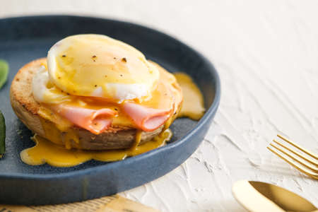 egg Benedict with hollandaise sauce on blue plate on table in white kitchenの写真素材