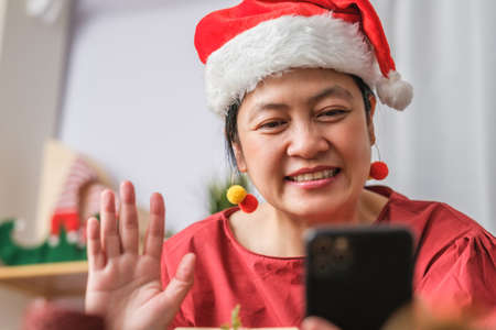 asian woman wear santa hat video call party with friends with mobile phone on table at homeの写真素材