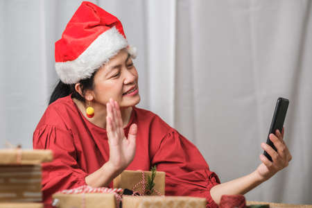 asian woman wear santa hat video call party with friends with mobile phone on table at homeの写真素材