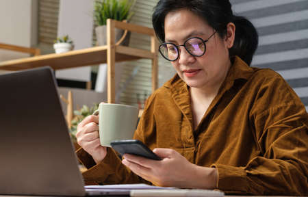 asian woman using mobile and drink coffee when take a break from work from laptop at homeの写真素材