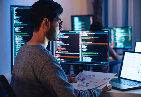 Asian software developers working on multiple screens displaying code and application diagrams at night in modern officeの写真素材