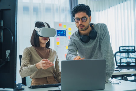 Asian software developer wearing a virtual reality headset works on a VR project, with a colleague focused on her computerの写真素材