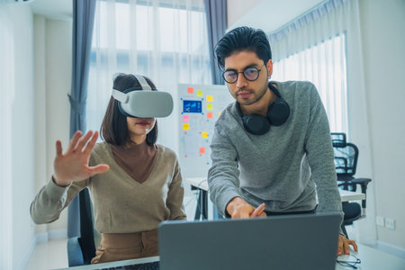 Asian software developer wearing a virtual reality headset works on a VR project, with a colleague focused on her computerの写真素材