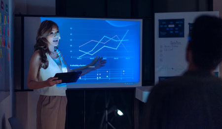 asian leader woman present  business presentation in a dimly lit office, analyzing financial charts and data projections on a screenの写真素材