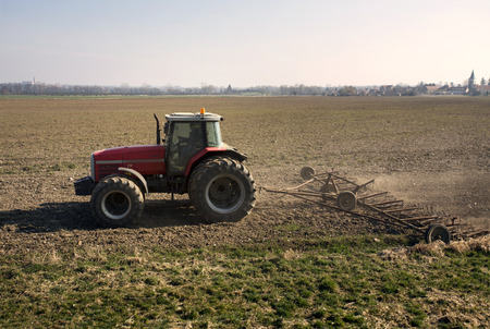 Red tractor works on field. Farmer harrows soil clods in rural area. Agriculture industrial motive with plow. Exterior profile countryside scene in spring. Graphic component, partition element.のeditorial素材