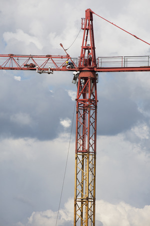 Assembling of crane on dramatic clouds background. Dangerous situation in height. Big yellow crane with a workman. High construction machinery with load, burden. Hook hangs on rope. Composition on Sky.の写真素材