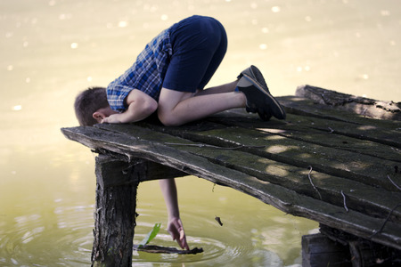 Thoughty boy on pier near pond and playing with small boat from bark and leaf. A little kid thinks and looks at the pond. Dream positive atmosphere, sunny day.の写真素材