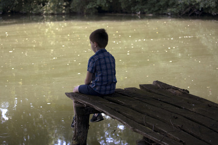 Dreamy boy sitting on a wooden pier at the pond. Little kid thinks and looks at the pond. Dream positive atmosphere, sunny day.の写真素材